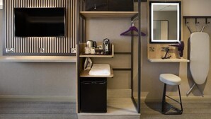 Habitación de hotel pequeña con televisión, un lavamanos, espejo, taburete y un mueble para guardar ropa colgada.