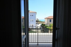 Une vue à travers une porte ouverte, donnant sur un balcon doté d’une rambarde en métal, et surplombant une rue bordée d’immeubles, avec un ciel dégagé.