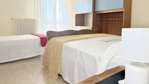 Et hotelværelse med to senge, et natbord, en lampe, et vindue med gardiner, en radiator og et klædeskab.
