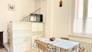 Et spiseområde med et bord dækket af en mønstret dug, stole, et skab med et fjernsyn ovenpå, og et vindue med gardiner.