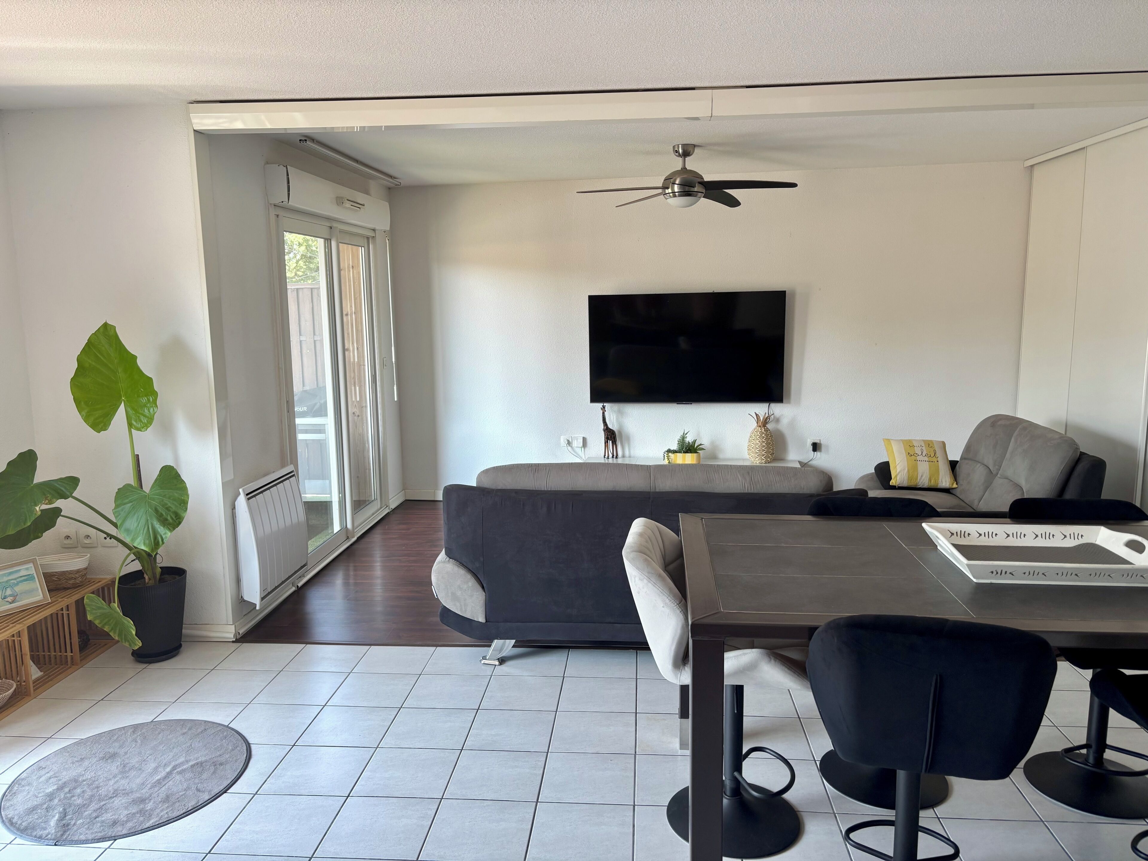 Una sala moderna con un sofá, una mesa de comedor con sillas, un ventilador de techo, un televisor de pantalla plana montado en la pared y una planta grande en maceta.
