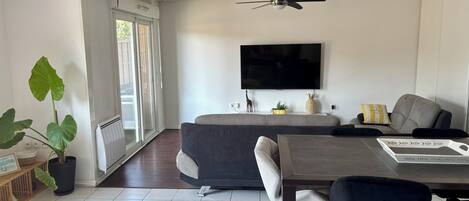 Una sala moderna con un sofá, una mesa de comedor con sillas, un ventilador de techo, un televisor de pantalla plana montado en la pared y una planta grande en maceta.
