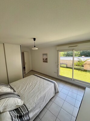 Ein Schlafzimmer mit Bett, Kleiderschrank, Tür, Deckenleuchte, Fenster und Blick ins Freie.