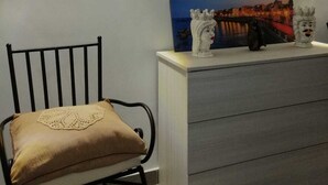 Ett rum med en svart metallstol, en beige kudde, en vit byrå och en väggmonterad lampa.
