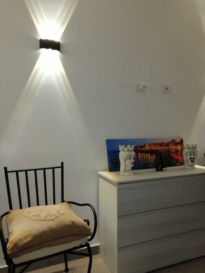 Ein Zimmer mit einem schwarzen Metallstuhl, einem beigen Kissen, einer weißen Kommode und einer wandmontierten Lampe.