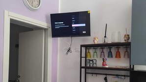 Ett rum med en tv som är monterad på väggen, en klocka, en hylla med diverse föremål och en dörr som leder till ett annat rum.