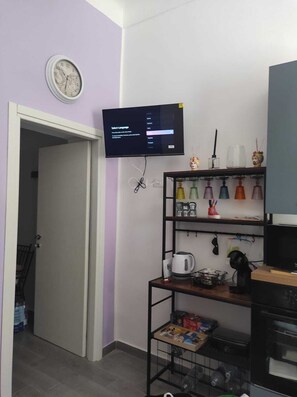 Ein Zimmer mit einem an der Wand montierten Fernseher, einer Uhr, einem Regal mit diversen Gegenständen und einer Tür zu einem anderen Raum.