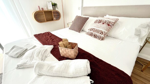 Una cama bien hecha con sábanas blancas, una manta roja y una cesta con artículos de aseo.