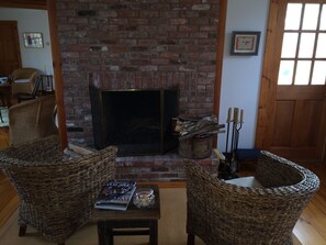 A living room with a brick fireplace, wicker chairs, a small table with a magazine, and a door with a window. - Gorgeous 3-bedroom house with AC in West Tisbury (West Tisbury)