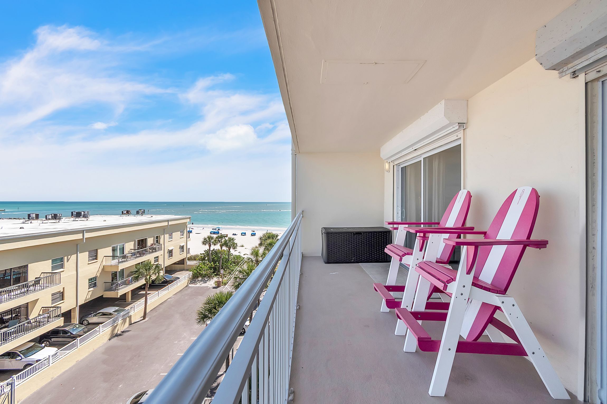 A balcony with a view of the beach, pink and white chairs, and a building with balconies and parked cars.