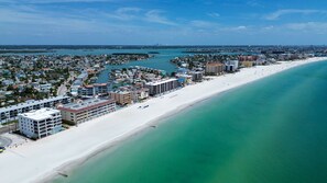Vue aérienne d’une ville côtière avec une longue plage de sable, un port de plaisance et plusieurs immeubles à étages.
