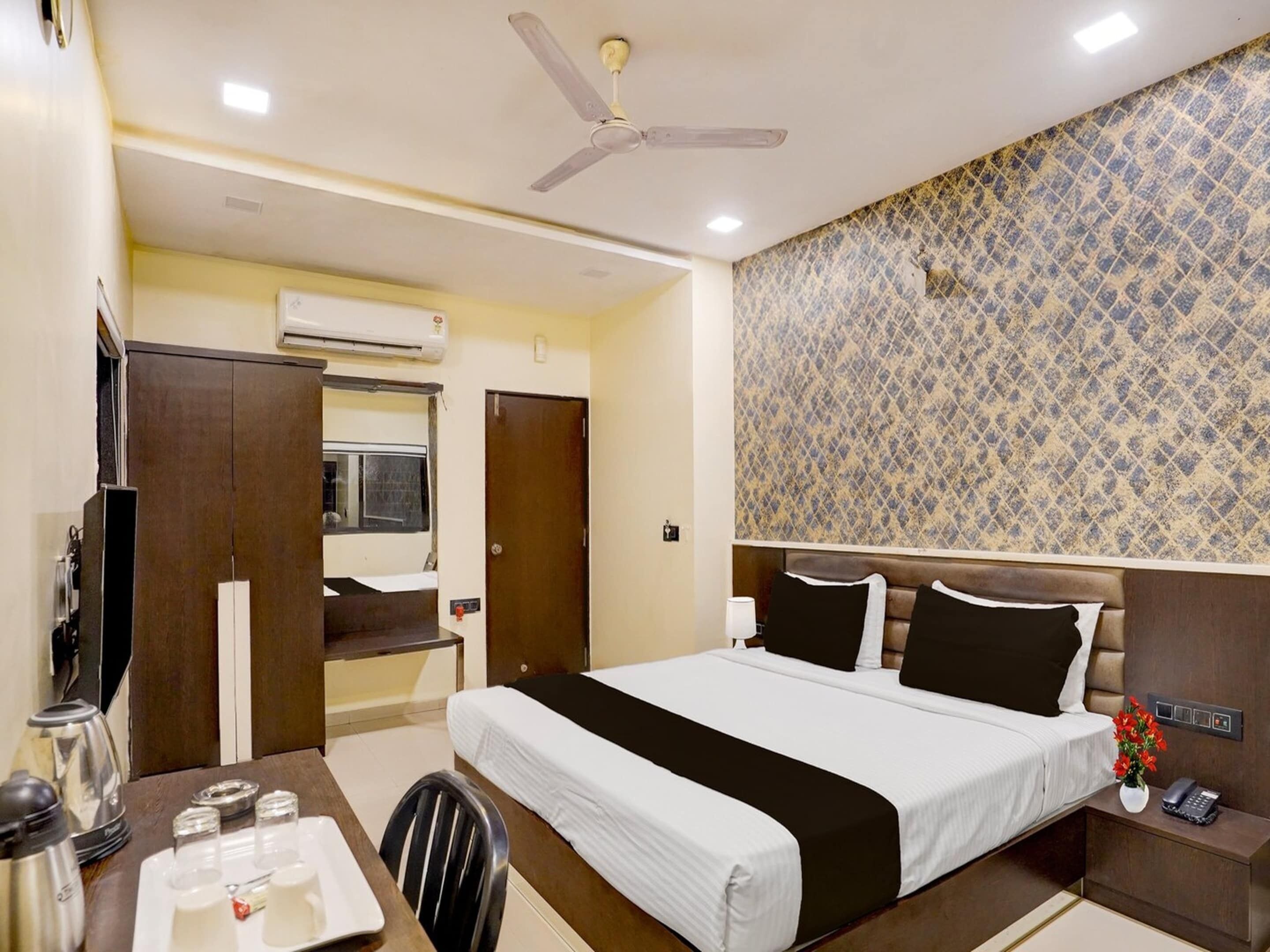 Une chambre d’hôtel avec un lit, un bureau et une chaise, une télévision, un ventilateur de plafond et un mur décoré de motifs.