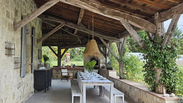 Ein Essbereich im Freien mit einer rustikalen Holz-Pergola, einem langen Tisch, der für eine Mahlzeit gedeckt ist, und einer Steinmauer mit Grünpflanzen.