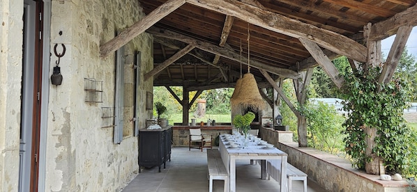 Ein Essbereich im Freien mit einer rustikalen Holz-Pergola, einem langen Tisch, der für eine Mahlzeit gedeckt ist, und einer Steinmauer mit Grünpflanzen.