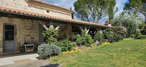 Ein Steinhaus mit Ziegeldach, einer überdachten Veranda und einem Rasen mit verschiedenen Sträuchern und Blumen.