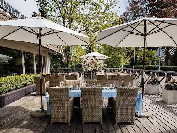 Un área de comedor al aire libre con sillas y mesas de mimbre, y sombrillas, ubicada en una terraza de madera con follaje de fondo.