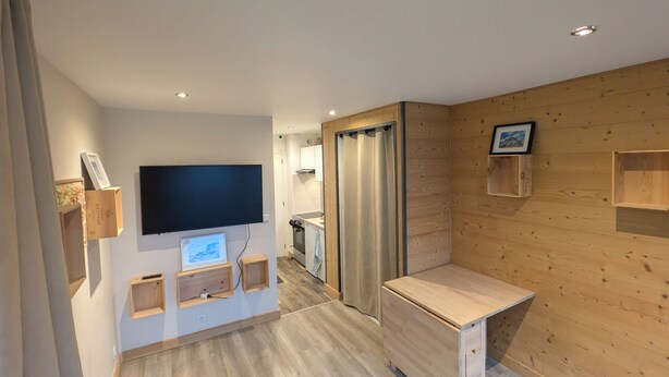 Ein modernes Zimmer mit Flachbildfernseher an der Wand, Holzschränken, einem Holztisch und einer Schiebetür zu einer Küchenzeile.