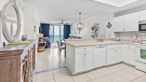 A modern kitchen with white cabinetry, a central island, and a dining area with blue curtains. - 🌞🩴Gulf-Front 3BR | Great Amenities + Steps to Beach | Summerwind West 1403 (Navarre Beach)