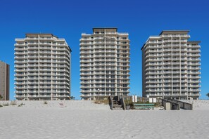 Drei Hochhäuser mit zahlreichen Balkonen, ein sandiger Strand im Vordergrund und ein klarer, blauer Himmel.