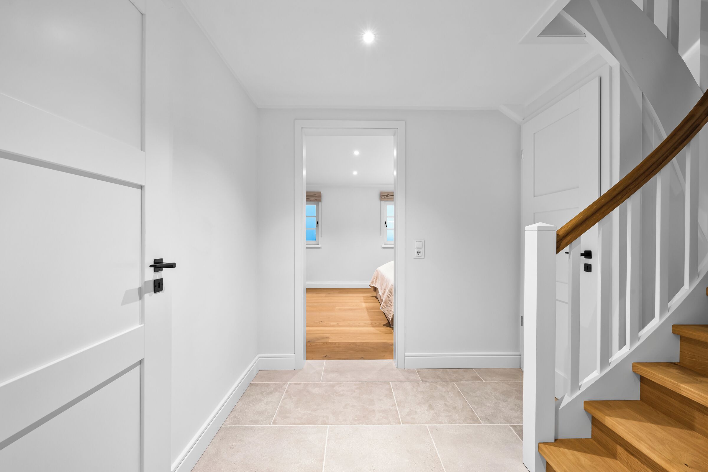A hallway with a wooden staircase, a door, and a view into a bedroom with a bed and a window.