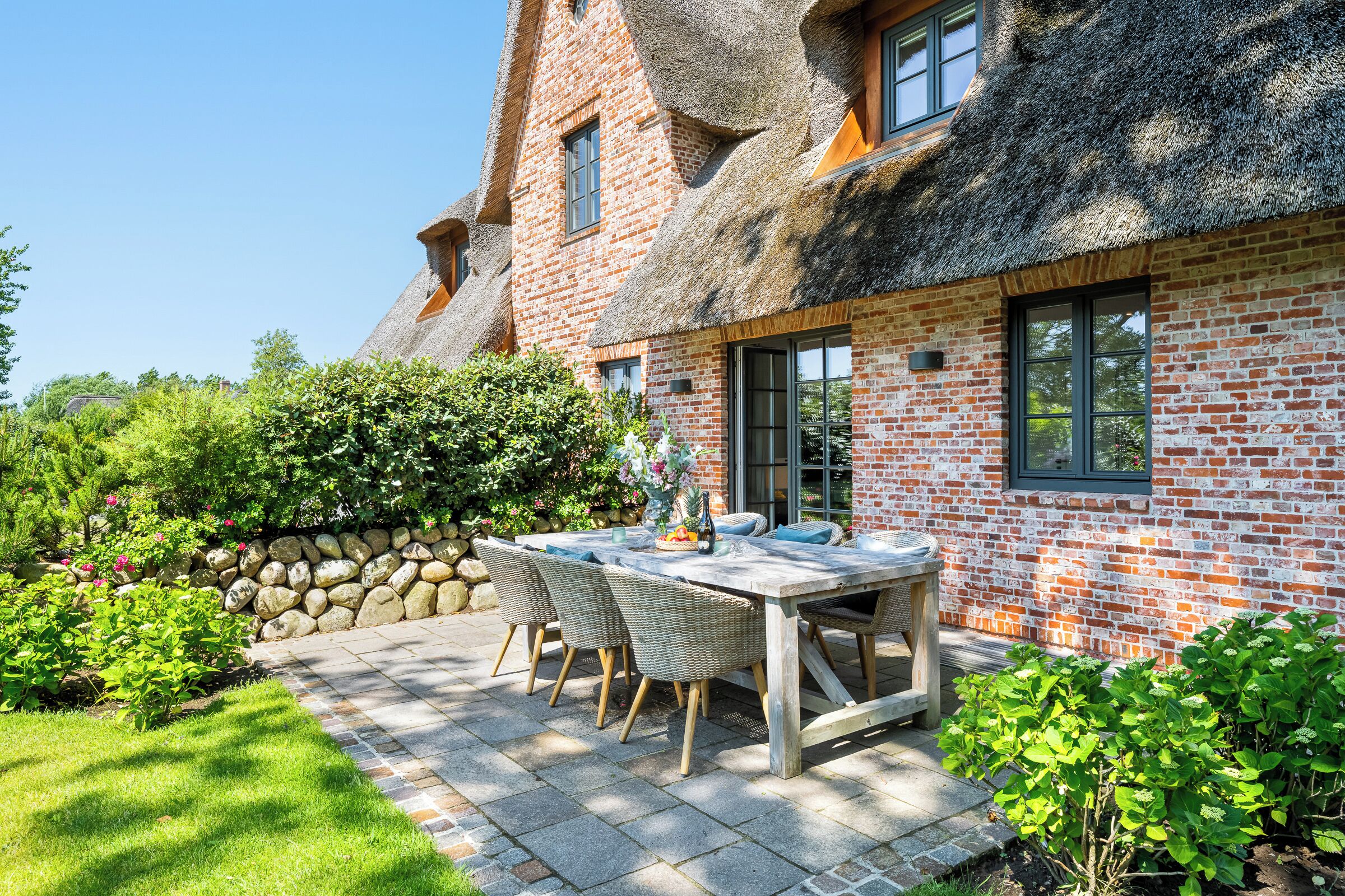 Une maison à toit de chaume, aux murs en briques, dotée d’une terrasse avec une table et des chaises, et d’un mur en pierre.