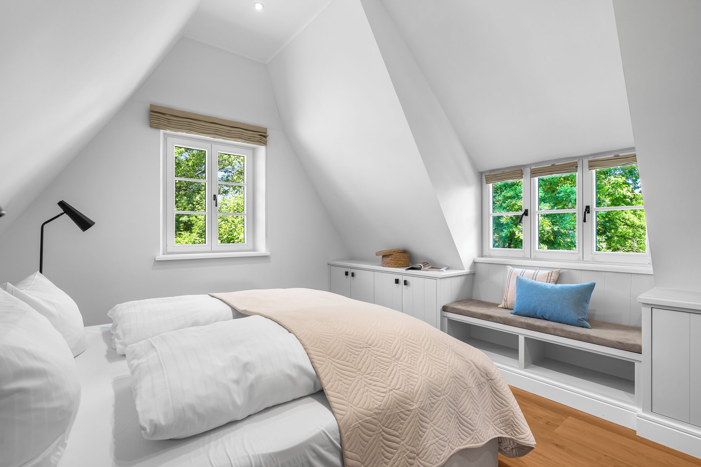Ein Schlafzimmer mit schräg gedachter Decke, einem Bett mit weißer Bettwäsche, einer Bank mit Kissen und zwei Fenstern mit weißen Rahmen.