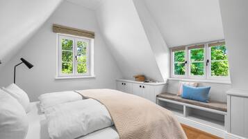 Ein Schlafzimmer mit schräg gedachter Decke, einem Bett mit weißer Bettwäsche, einer Bank mit Kissen und zwei Fenstern mit weißen Rahmen.
