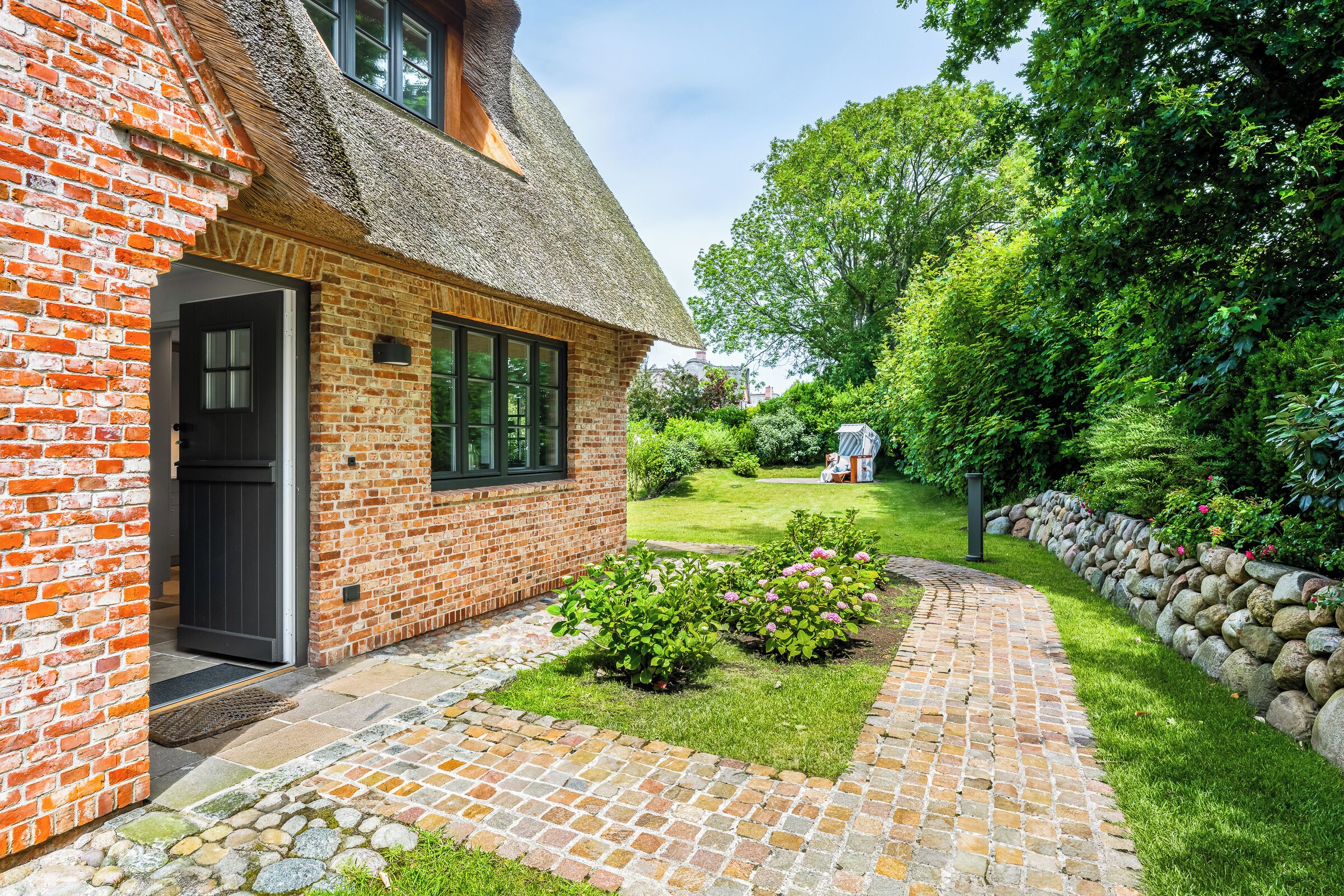 A brick house with a thatched roof, a paved garden pathway, and a stone wall.
