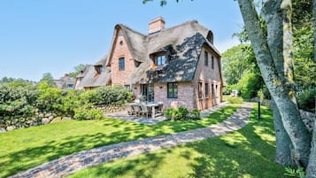 A large brick house with a thatched roof, surrounded by a well-maintained garden and a stone pathway.