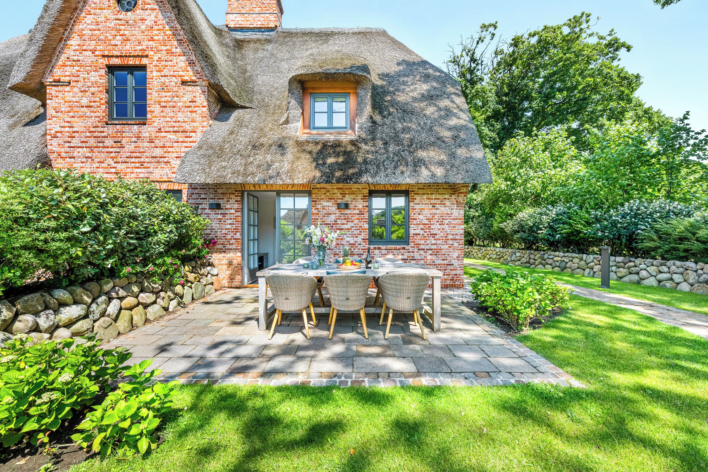 A brick house with a thatched roof, a patio with a table and chairs, and a stone wall.
