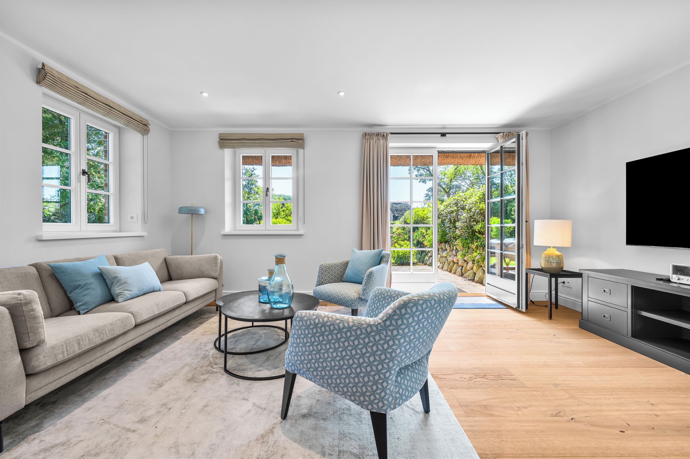 Un salon moderne comprenant un canapé, un fauteuil et une table basse, un téléviseur, et offrant une vue sur la verdure grâce à de grandes fenêtres.