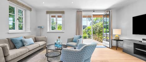 Un salon moderne comprenant un canapé, un fauteuil et une table basse, un téléviseur, et offrant une vue sur la verdure grâce à de grandes fenêtres.