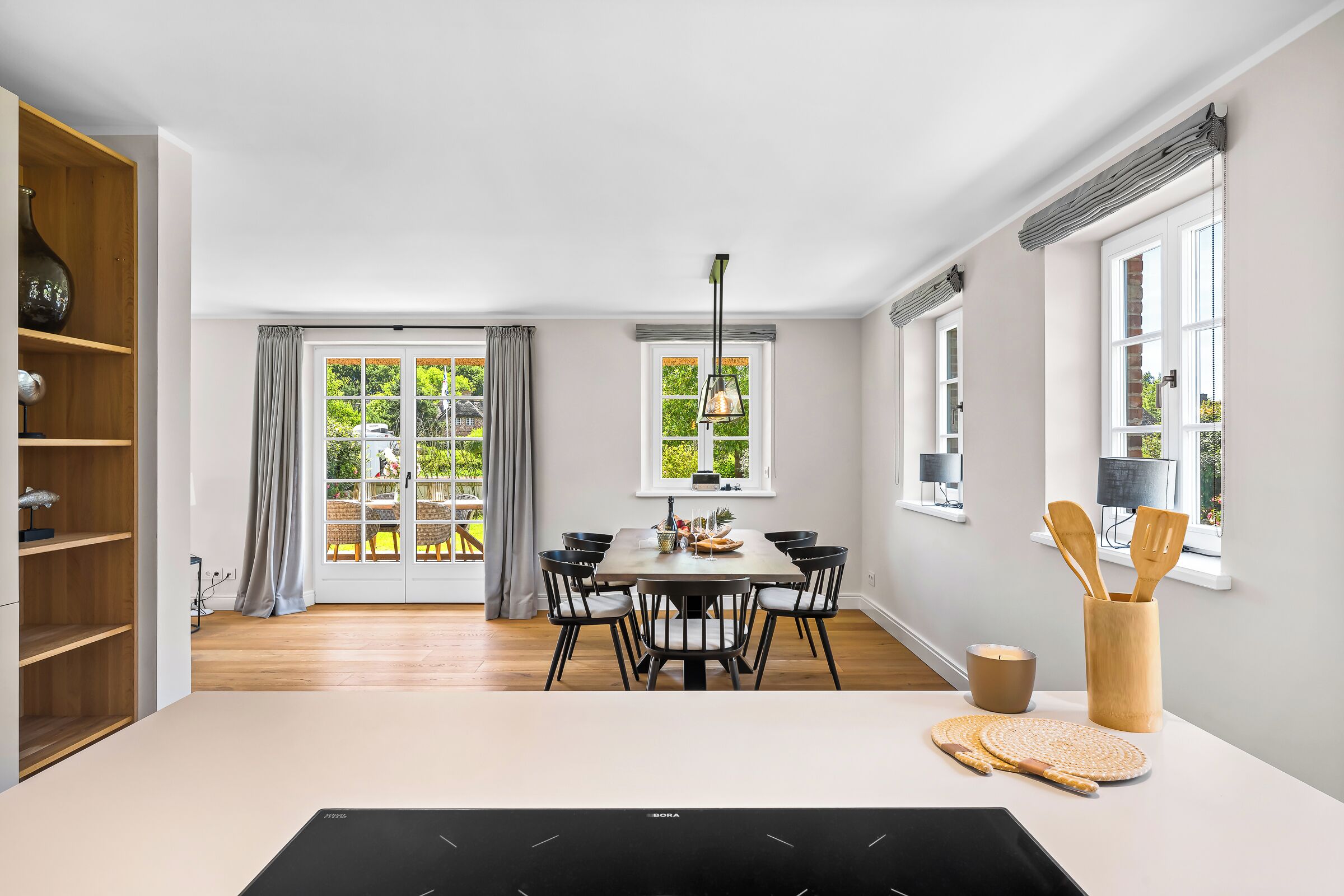 Et moderne spiseområde med et bord dækket til seks personer, et træskab og udsigt til grønne omgivelser.