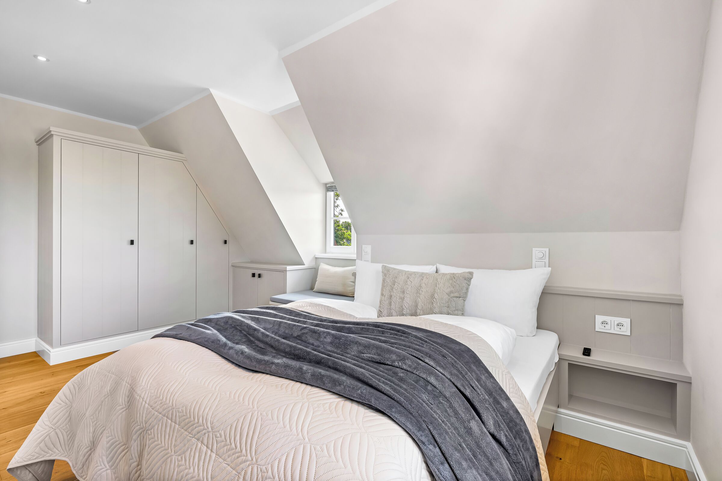 Un dormitorio con techo inclinado, una cama con manta gris, ropa de cama blanca y un armario blanco.
