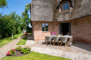 Ein Haus aus Ziegelsteinen mit einem Strohdach, einer Terrasse mit Tisch und Stühlen sowie einem Garten mit Blumen.