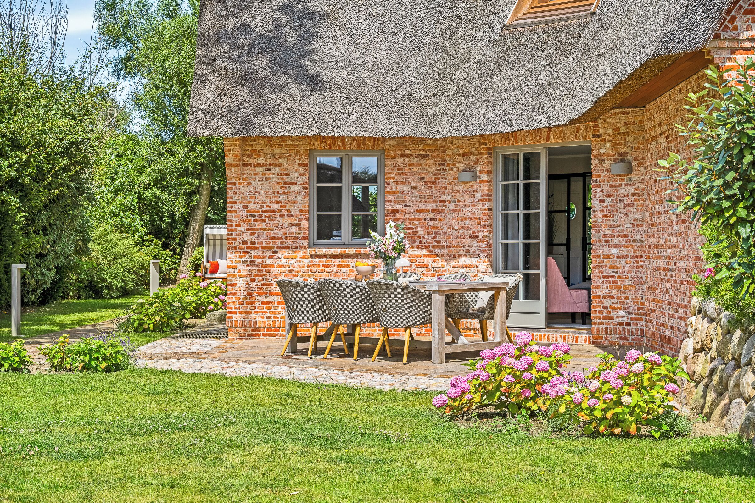 Ein Backsteinhaus mit einem Strohdach, einer Terrasse mit Tisch und Stühlen sowie ein Garten mit rosa Blüten.
