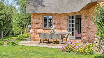 Ein Backsteinhaus mit einem Strohdach, einer Terrasse mit Tisch und Stühlen sowie ein Garten mit rosa Blüten.