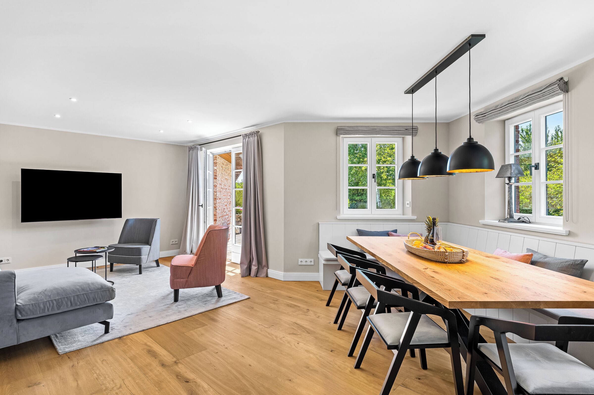 Un salon moderne doté d’un téléviseur à écran plat, de canapés gris, d’une table à manger en bois avec des chaises, et d’un coin cuisine avec une fenêtre donnant sur un espace verdoyant.
