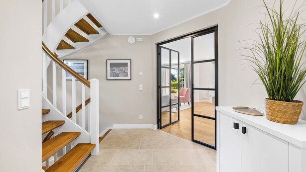 Um interior moderno com escada de madeira, parede branca com quadro emoldurado, porta de vidro deslizante para área externa e planta em vaso sobre um armário branco.