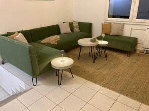 A green sofa with a throw blanket, two round tables with lamps, and a window with blinds. - The Corniche and its panoramic view (Fécamp)