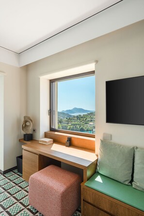 Ein Zimmer mit Schreibtisch, einem grünen Sofa, einem Fenster mit Aussicht und einem Wandfernseher.