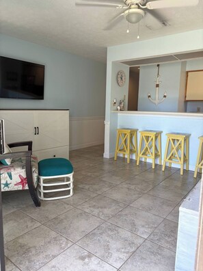 A living room with a TV, a white cabinet, a teal cushioned stool, and a bar area with stools. - Welcome to Margaritaville, Where it’s always 5 o’clock! Steps from the beach! (Panama City Beach)