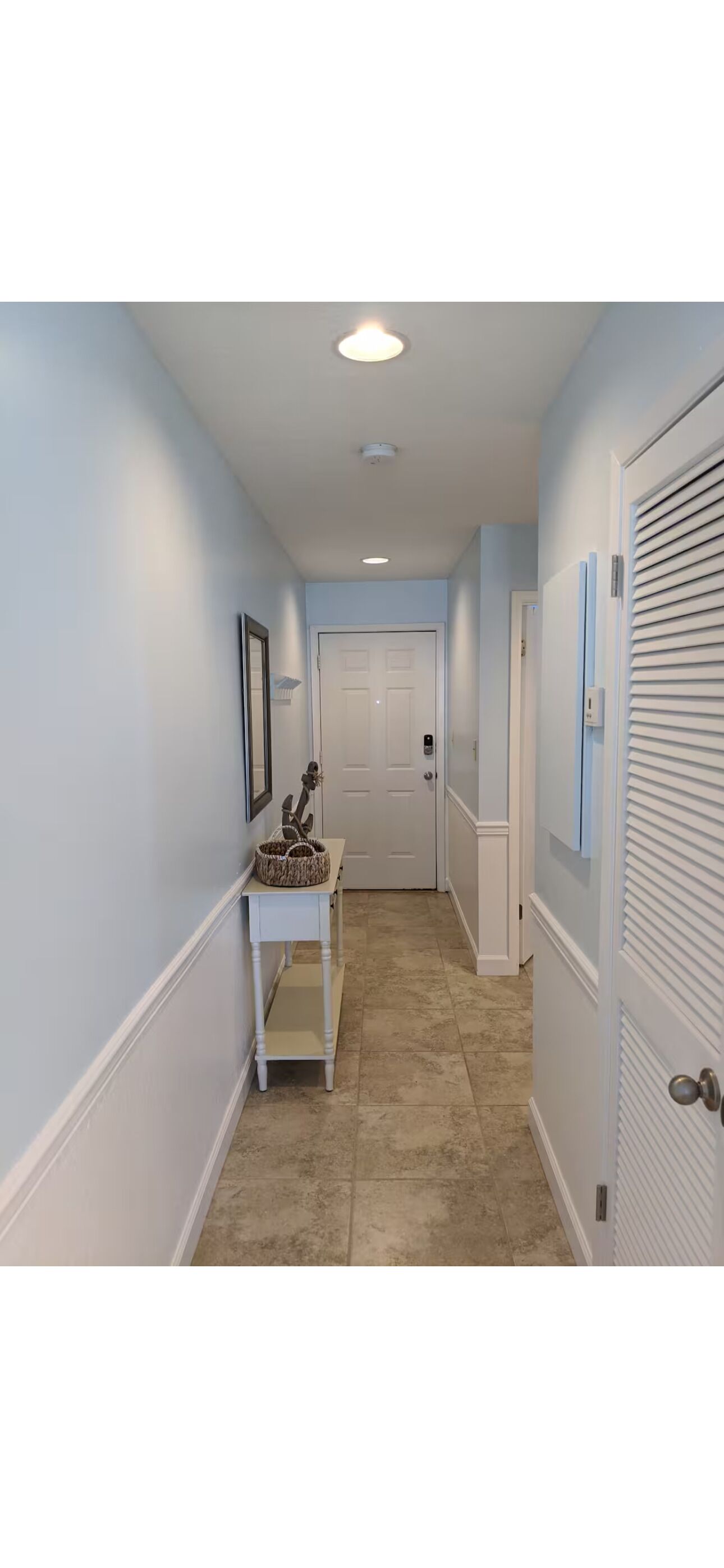 A hallway with a white door at the end, a small table with a basket, and a wall-mounted picture frame.