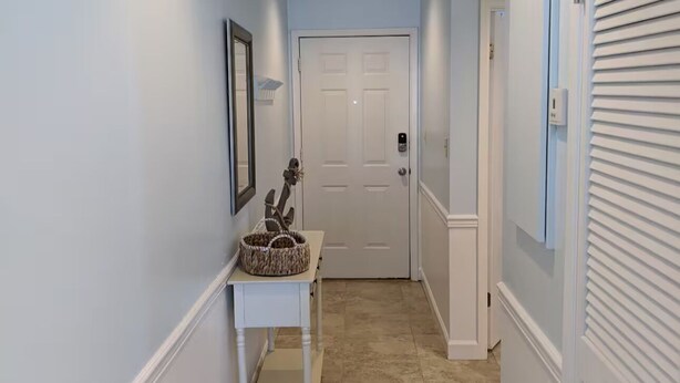 Un couloir avec une porte blanche au fond, une petite table sur laquelle se trouve un panier, et un cadre photo fixé au mur.