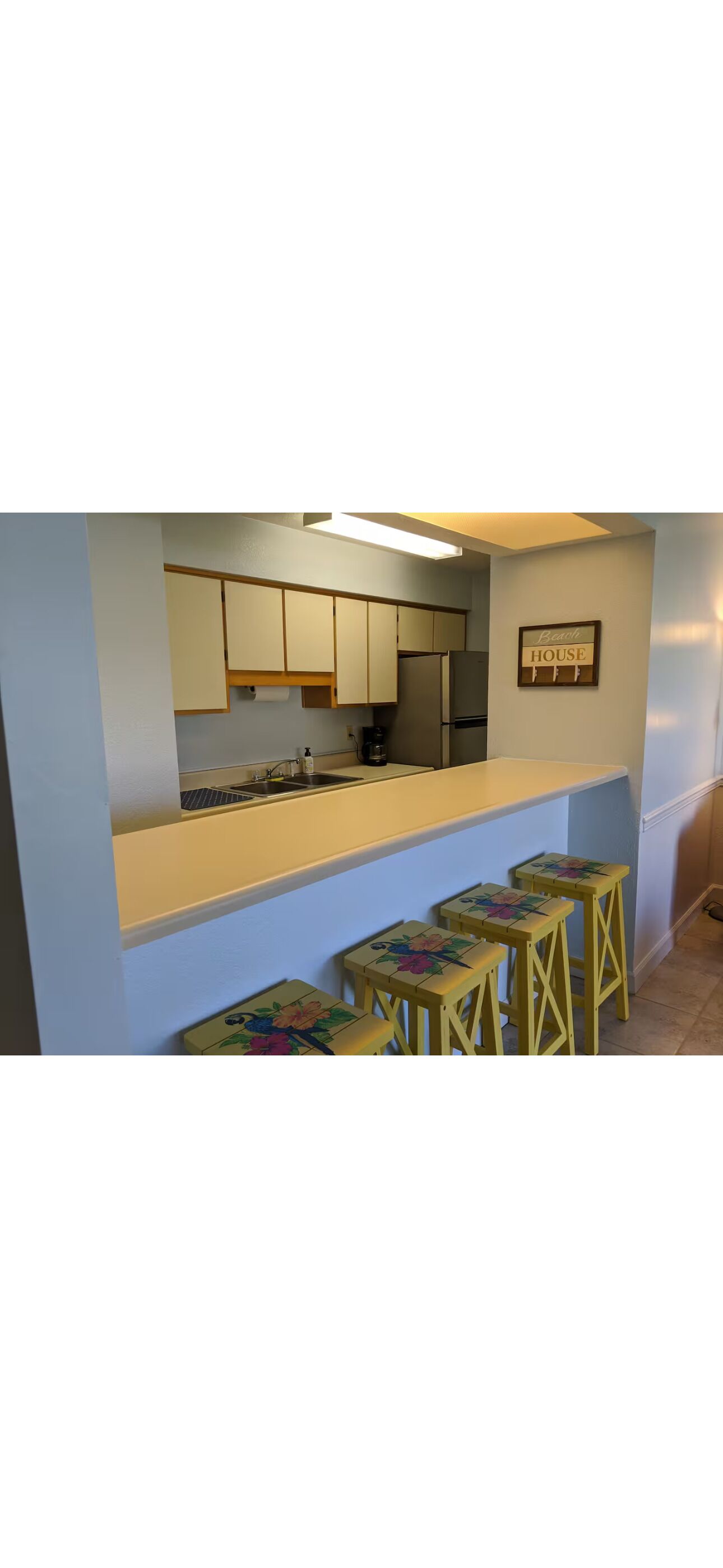 A kitchen with a countertop, cabinets, a refrigerator, and a sign that reads 'Beau House.' There are three stools with floral designs.