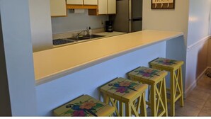 Une cuisine équipée d’un plan de travail, de placards, d’un réfrigérateur et d’une enseigne sur laquelle on peut lire « Beau House ». On y trouve également trois tabourets ornés de motifs floraux.