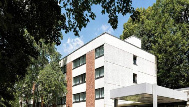 Een modern gebouw met een overdekte ingang, grote ramen en een parkeerplaats.