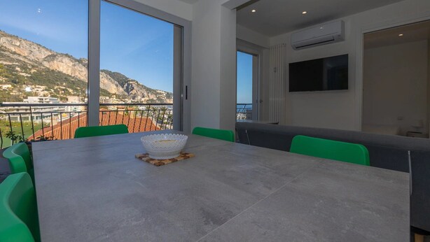 Ein moderner Essbereich mit einem großen Tisch, grünen Stühlen und Blick auf eine gebirgige Landschaft durch große Fenster.