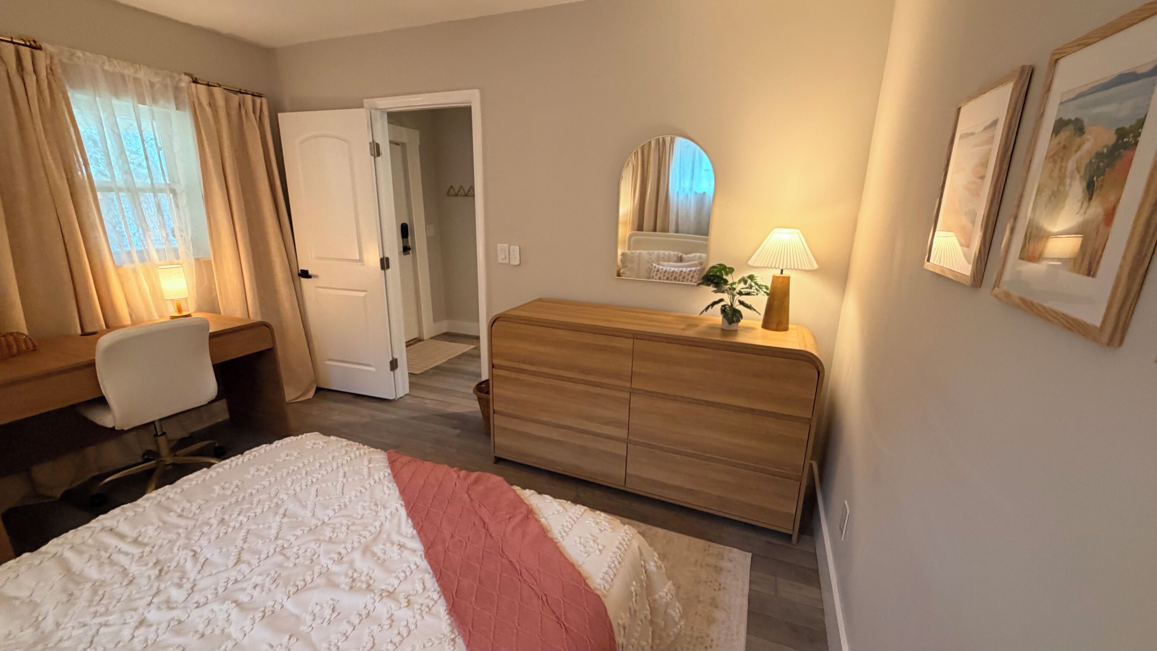 A bedroom with a bed, a desk, a dresser, a mirror, and a door leading to another room.