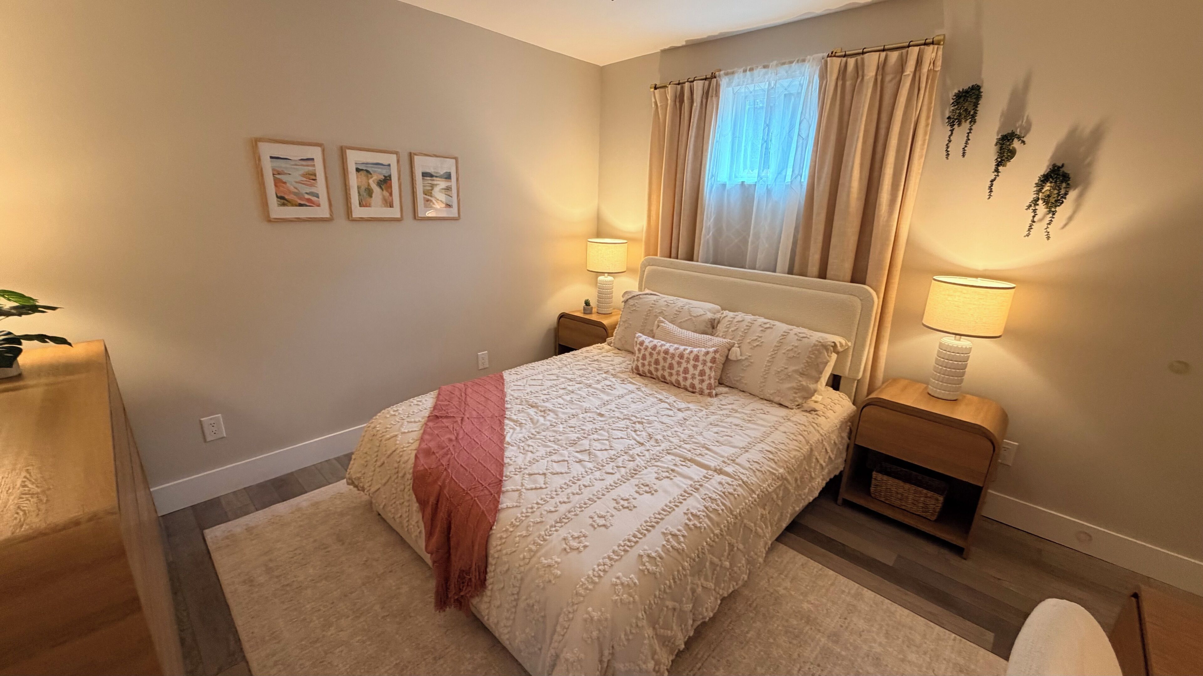 Une chambre à coucher avec un lit, deux tables de chevet, une commode, une lampe, une plante et trois tableaux encadrés accrochés au mur.